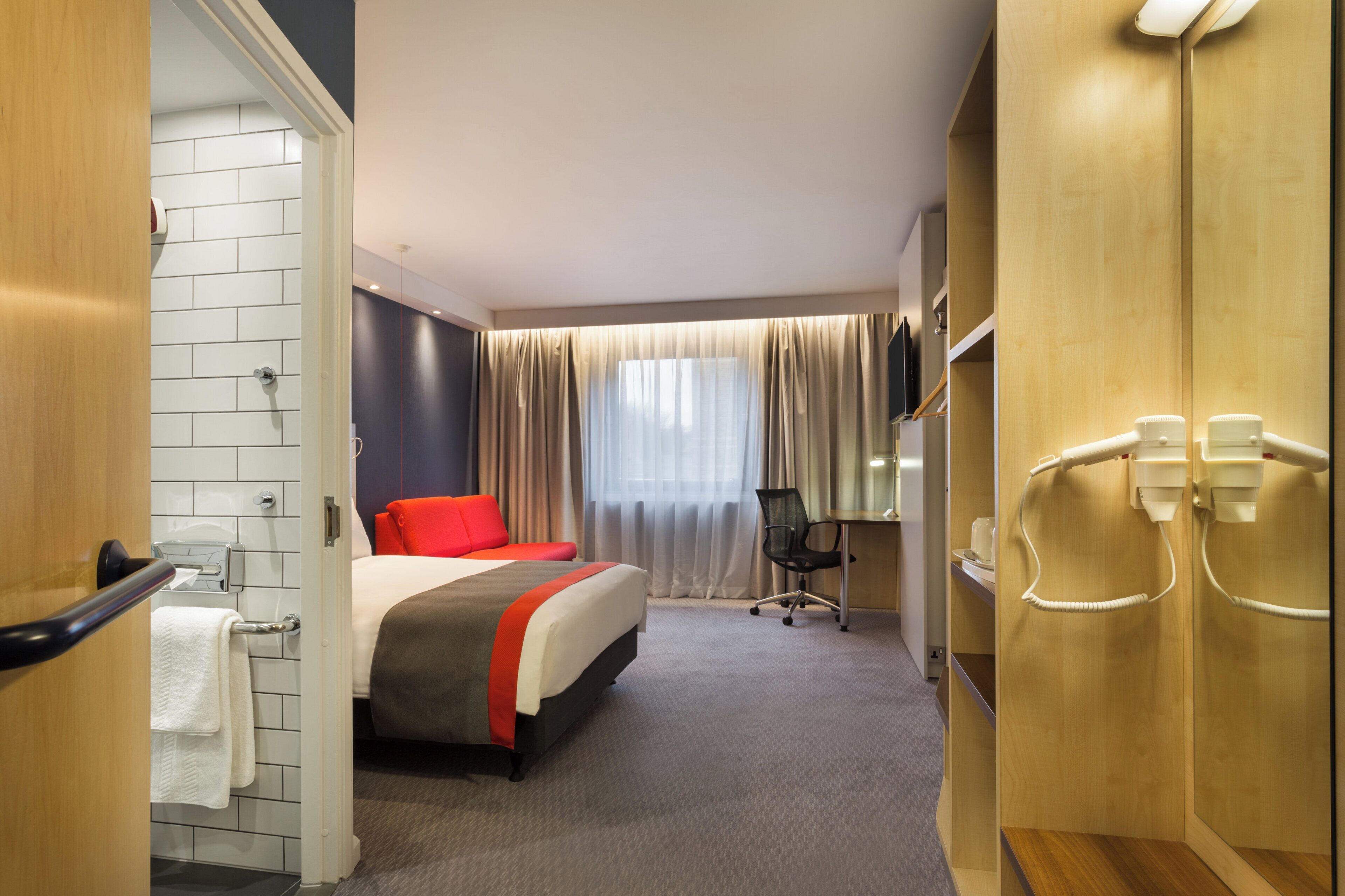 Habitación de hotel con cama, escritorio, silla, zona de ducha y televisor.