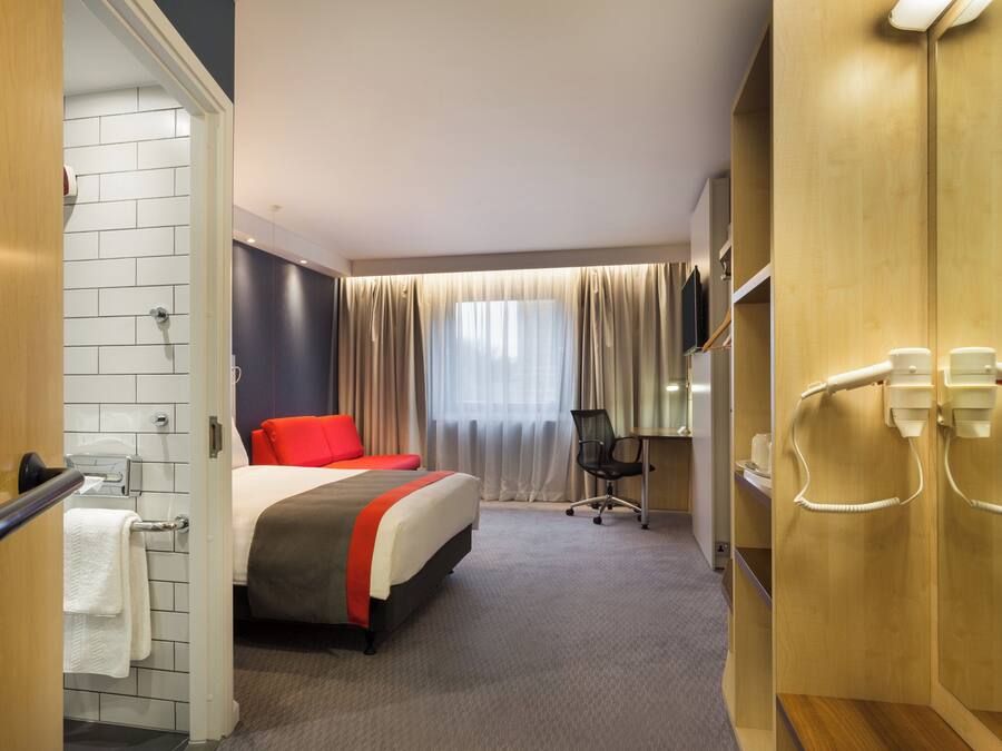 Habitación de hotel con cama, escritorio, silla, zona de ducha y televisor.