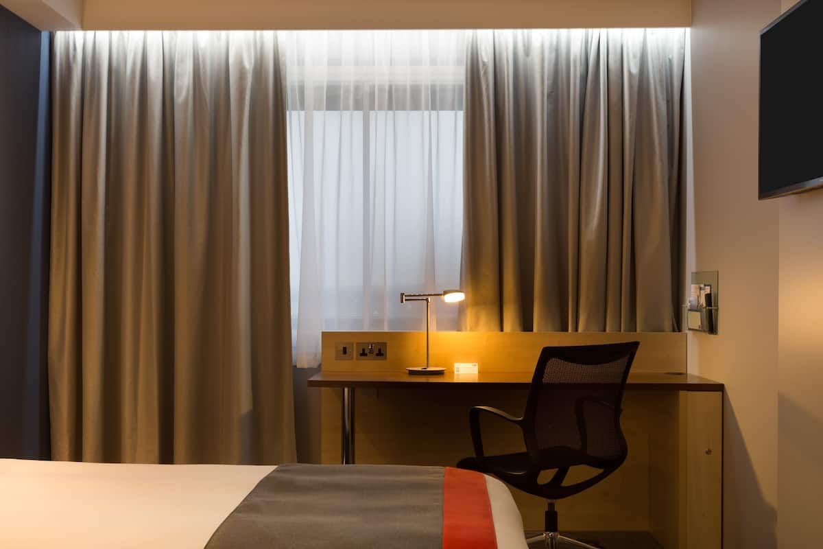 A hotel room with a bed, a desk with a chair, a desk lamp, and a window with curtains.