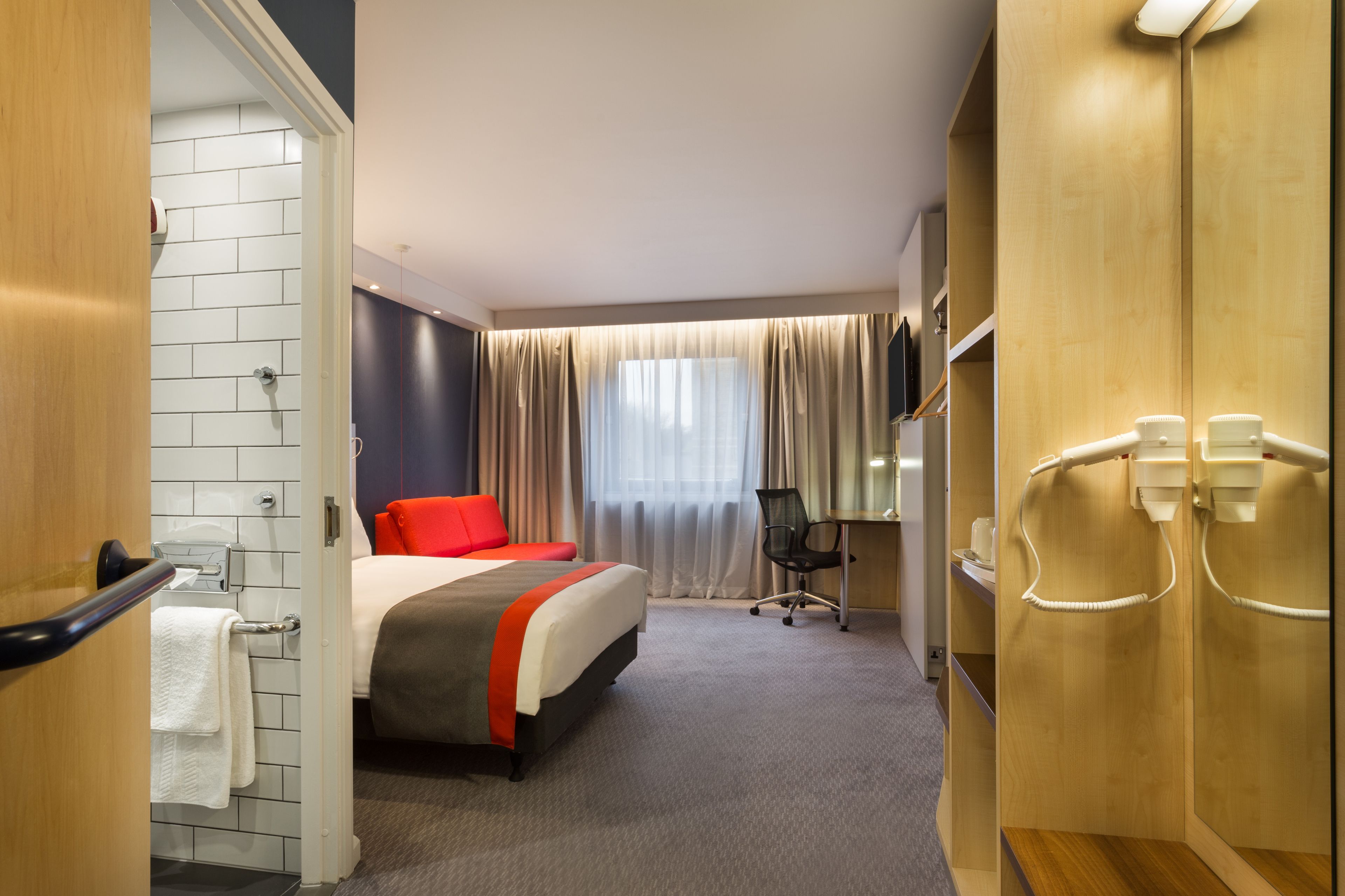 A hotel room with a bed, a desk, a chair, a mirror, and a bathroom with a towel rack.