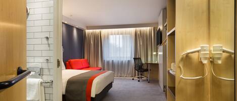 A hotel room with a bed, a desk, a chair, a mirror, and a bathroom with a towel rack.