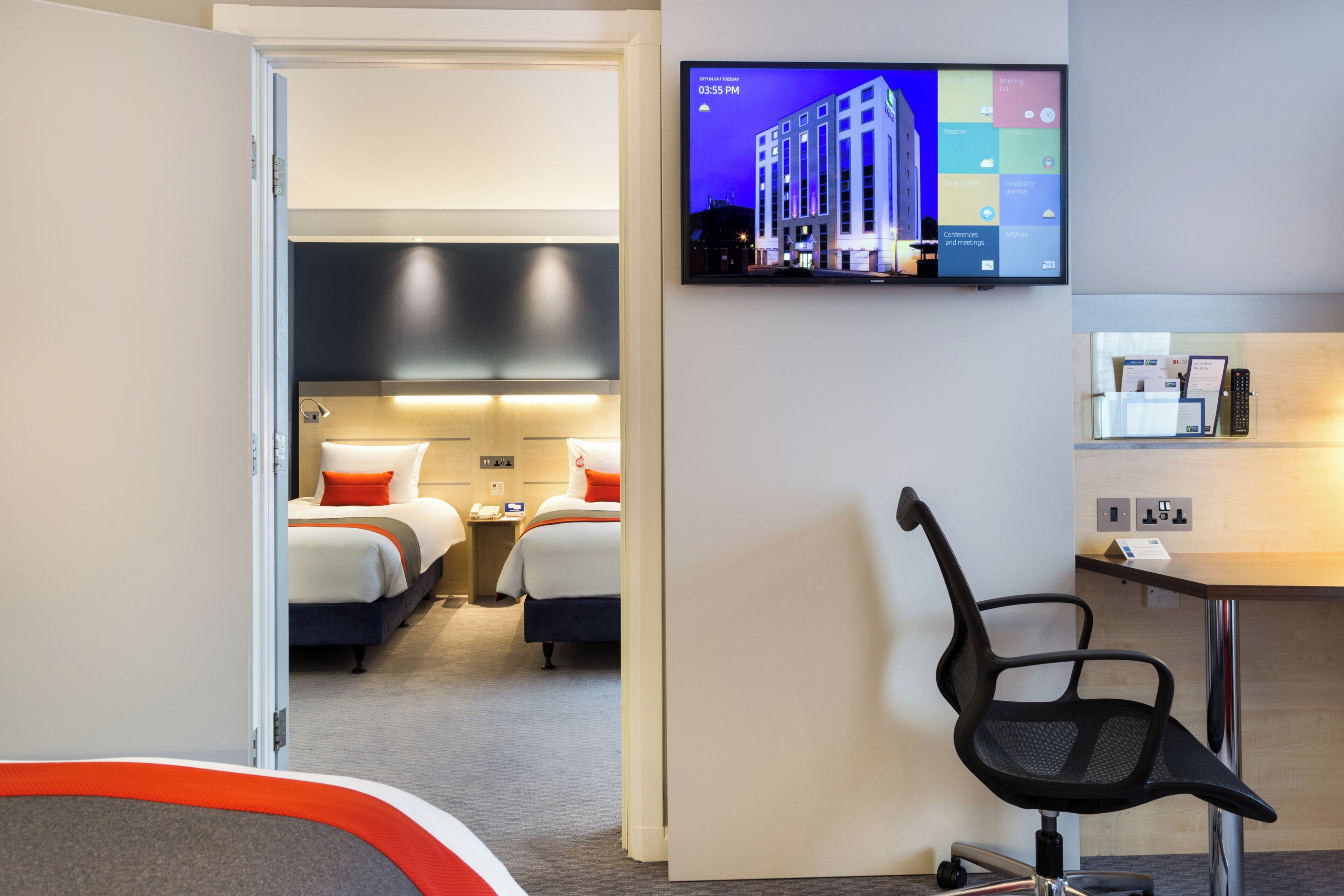 A hotel room with a bed, bedside table, lamp, and a TV mounted on the wall displaying a hotel building.