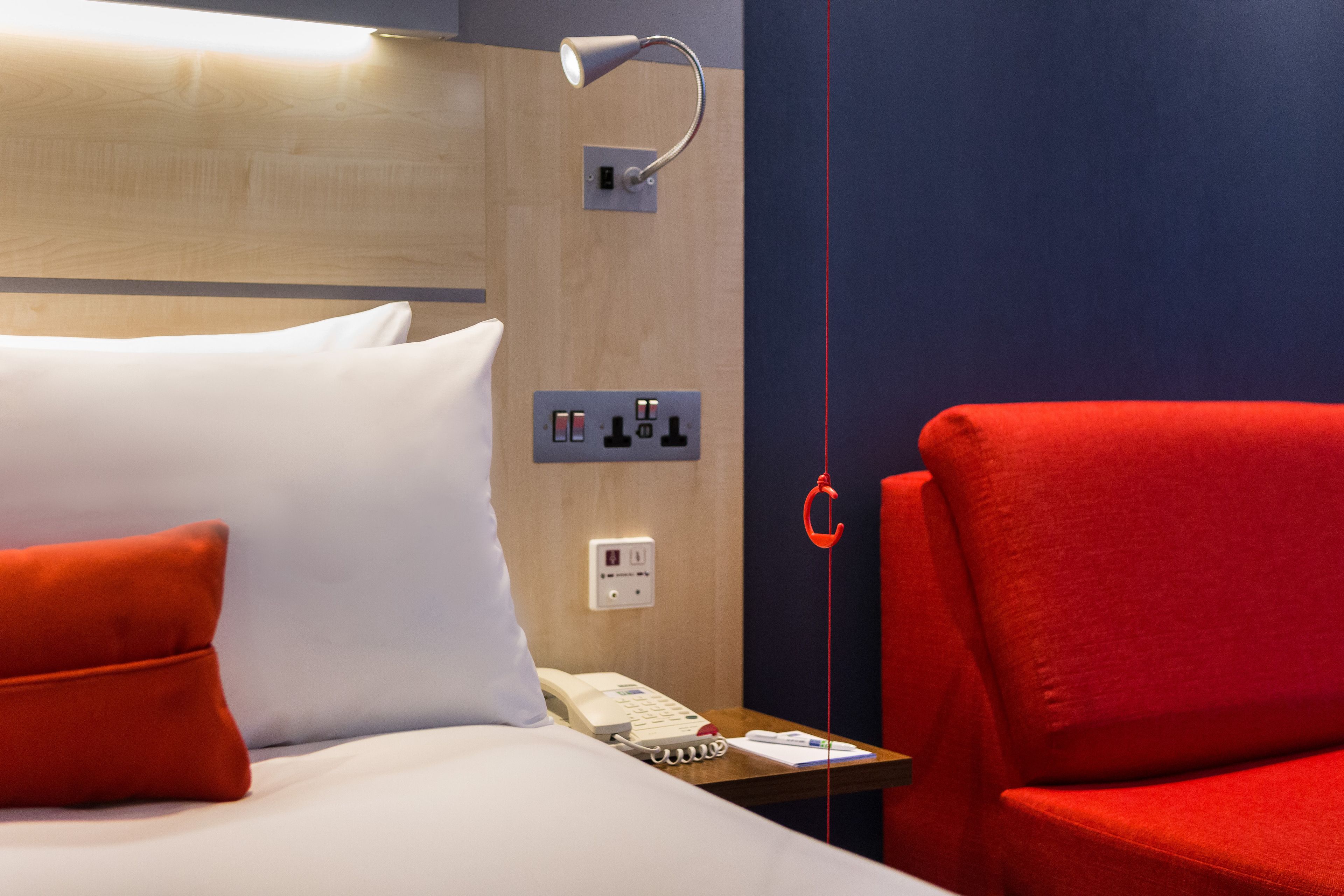 A hotel room with a bed, a red chair, a bedside table with a phone, and a wall-mounted light fixture.