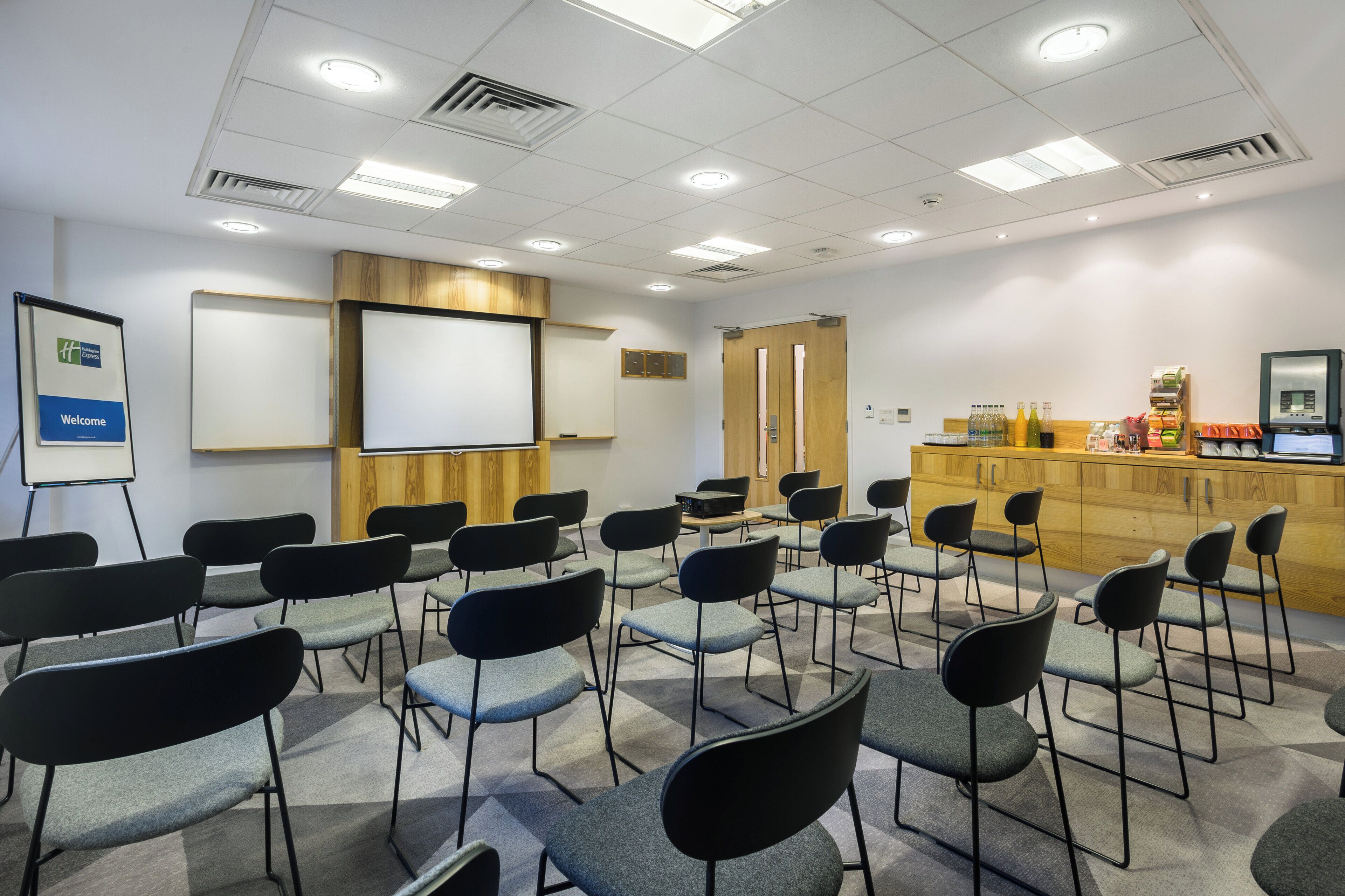 A conference room with rows of chairs, a whiteboard, a projector, and a wooden counter with beverages.