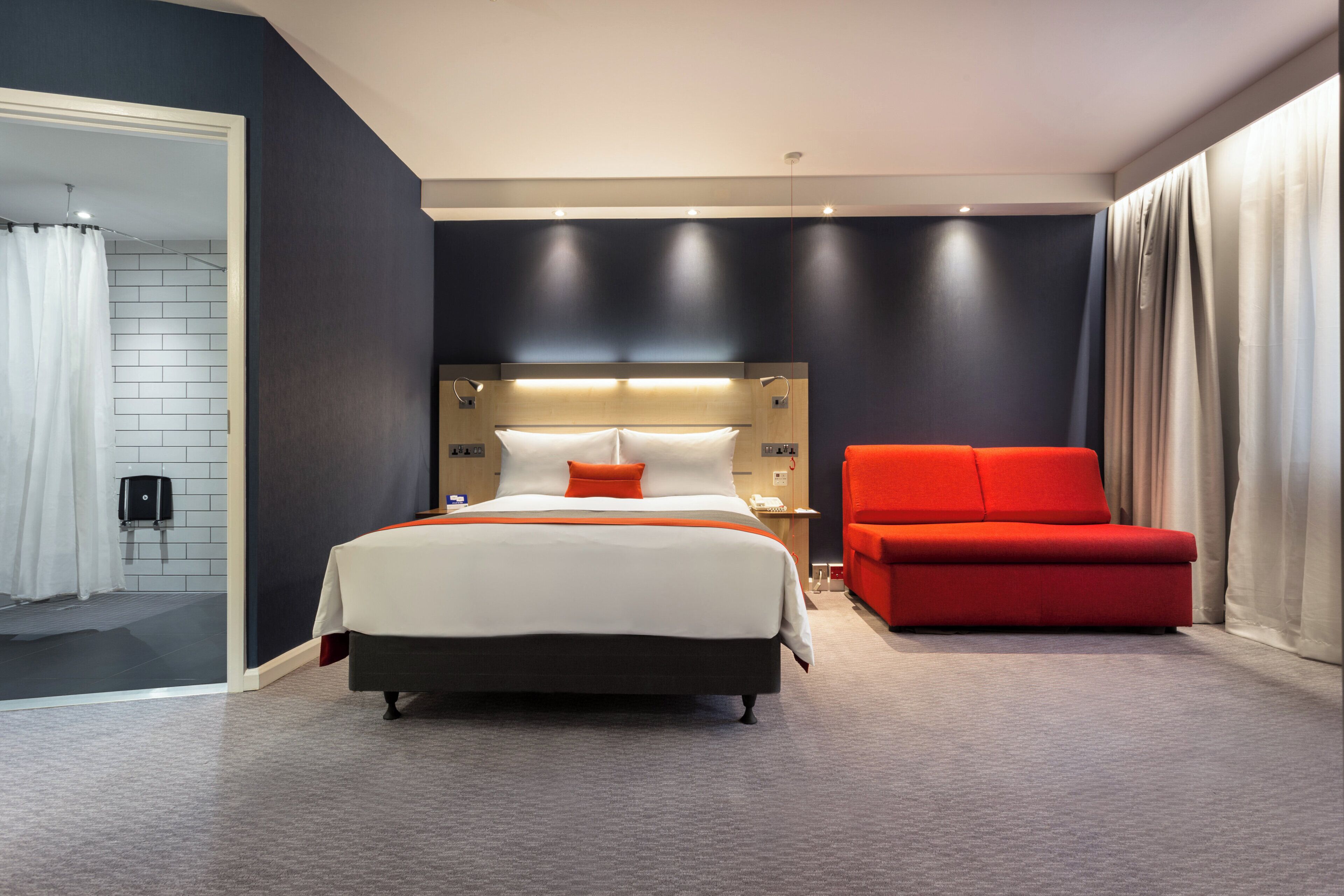 Une chambre d’hôtel moderne avec un grand lit, un fauteuil rouge et une salle de bain avec un rideau de douche blanc.