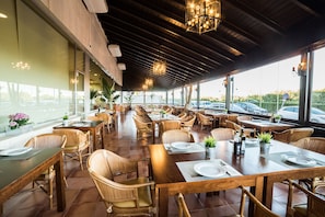 Un restaurante con mesas y sillas de madera, ventanas amplias y vistas a coches aparcados afuera.
