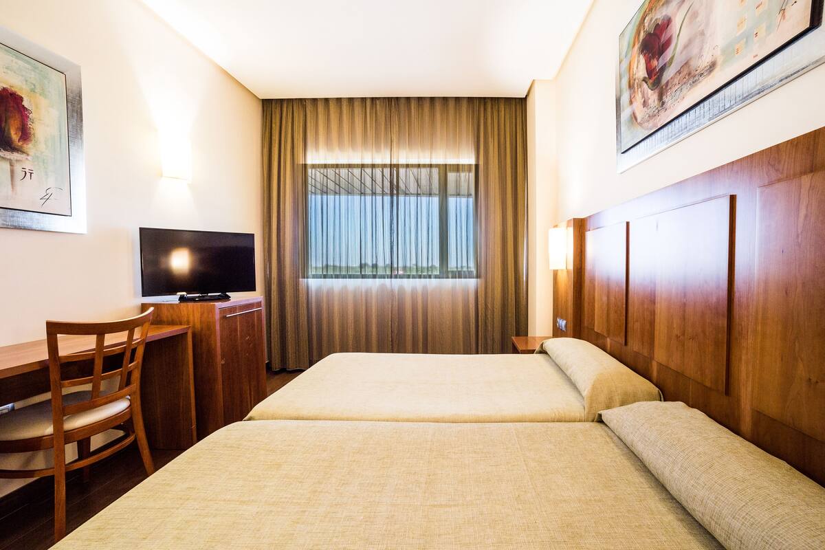 Habitación de hotel con dos camas, un escritorio con silla, televisor y un ventanal con cortinas.