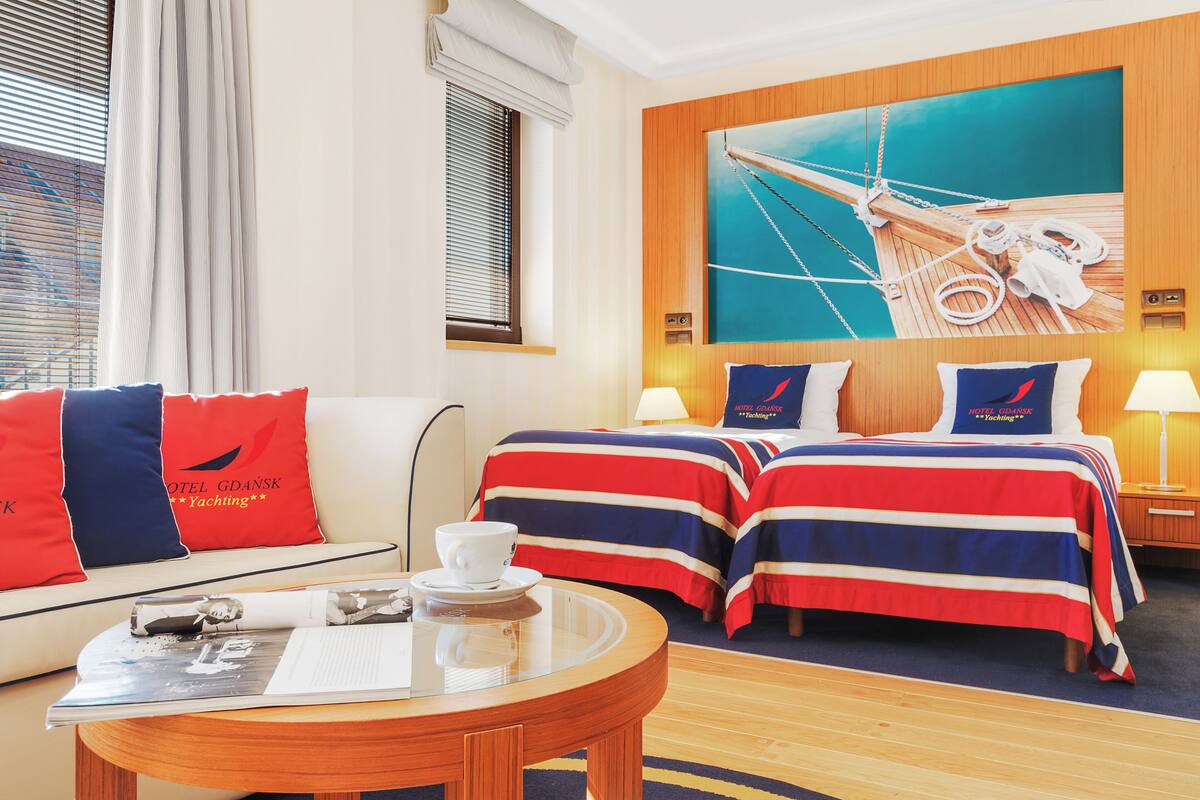A hotel room with two beds, a sofa, a round table, and a large painting of a ship's deck.
