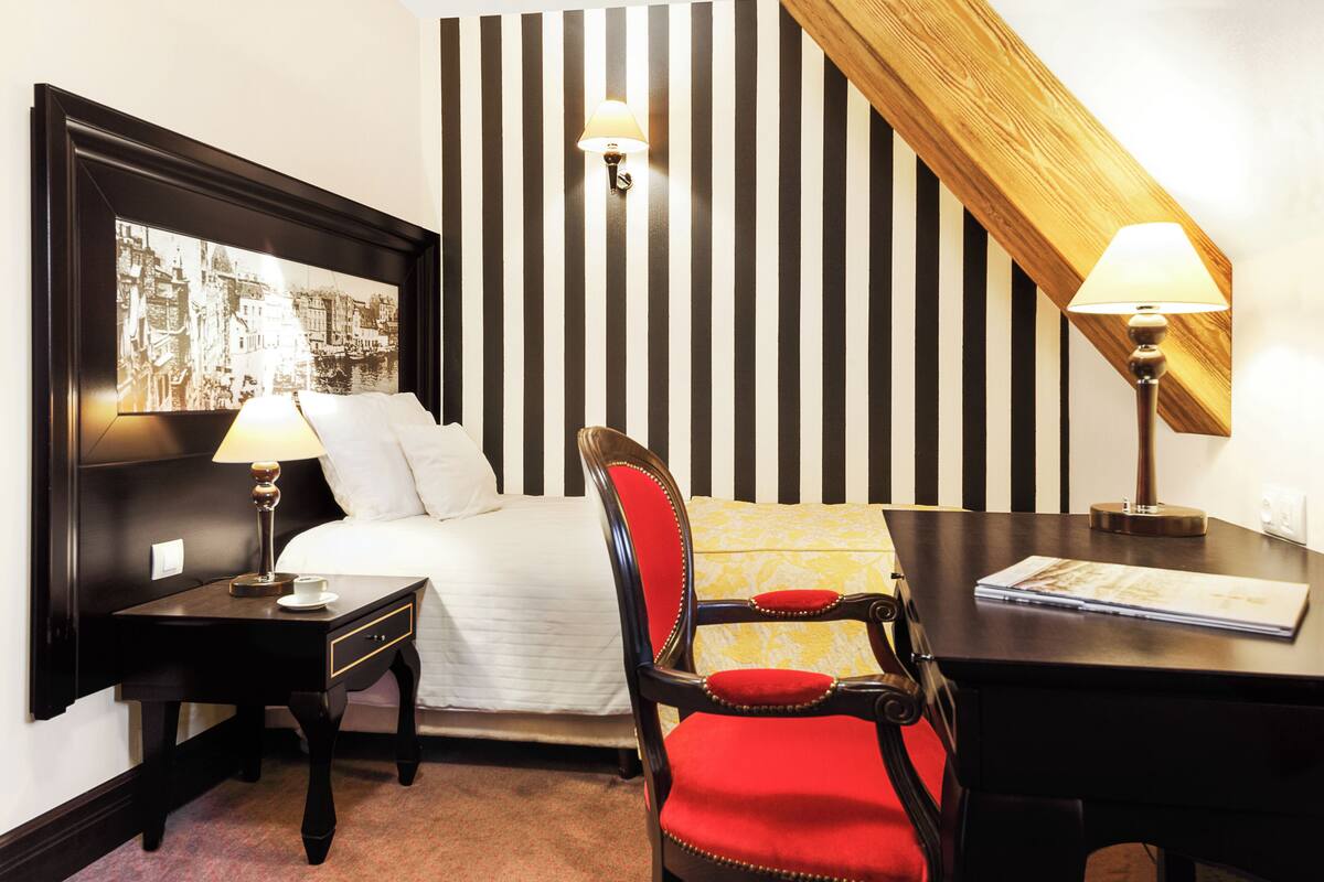 A hotel room with a bed, a desk, a chair, a nightstand, a lamp, and a wall-mounted light fixture.