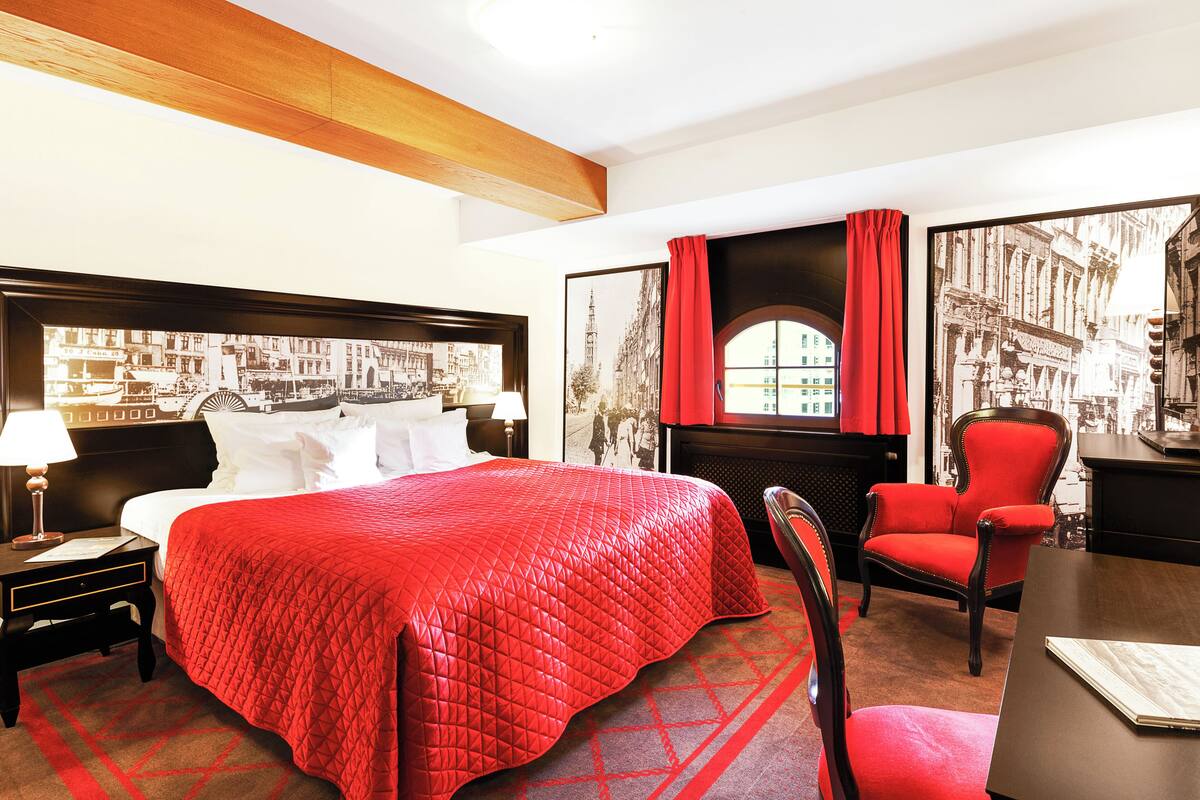 A hotel room with a large bed, a desk, a chair, and two large framed pictures on the wall.
