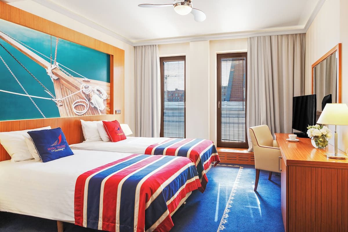 A hotel room with two beds, a desk, a chair, a television, and a large window with curtains.