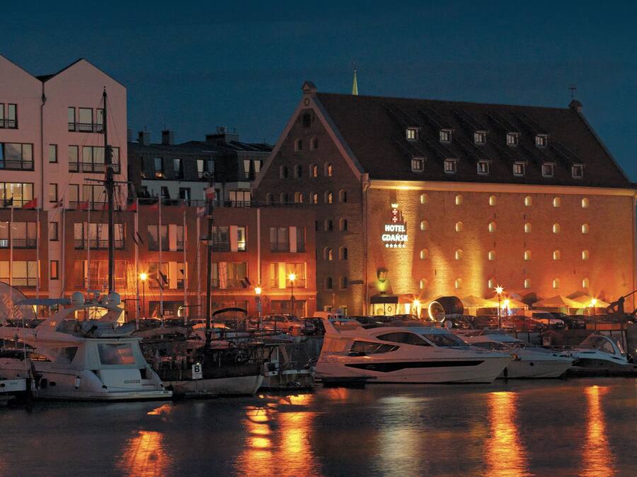 A marina with several yachts docked, a hotel with a prominent sign, and buildings with balconies and windows.