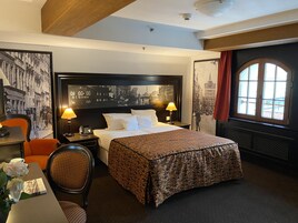 Una camera d'albergo con un letto grande, due abat-jour, una sedia, un tavolino con un telefono e una tazza, e una finestra con tende.