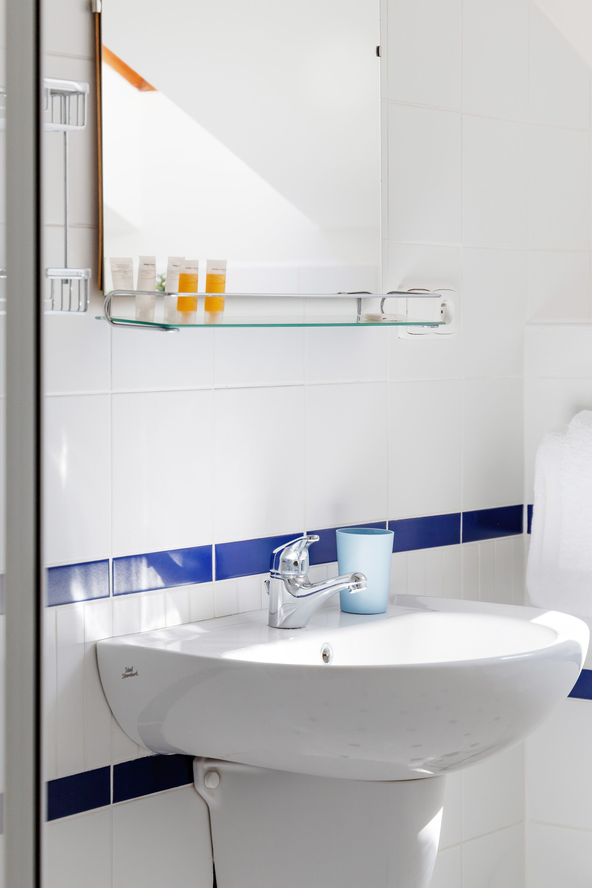 Un baño con un lavamanos blanco, azulejos azulados a rayas, un estante de vidrio con artículos de aseo personal y un espejo.