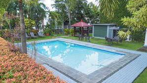 Piscina rectangular con solárium alicatado, rodeada de césped, palmeras y plantas en macetas.