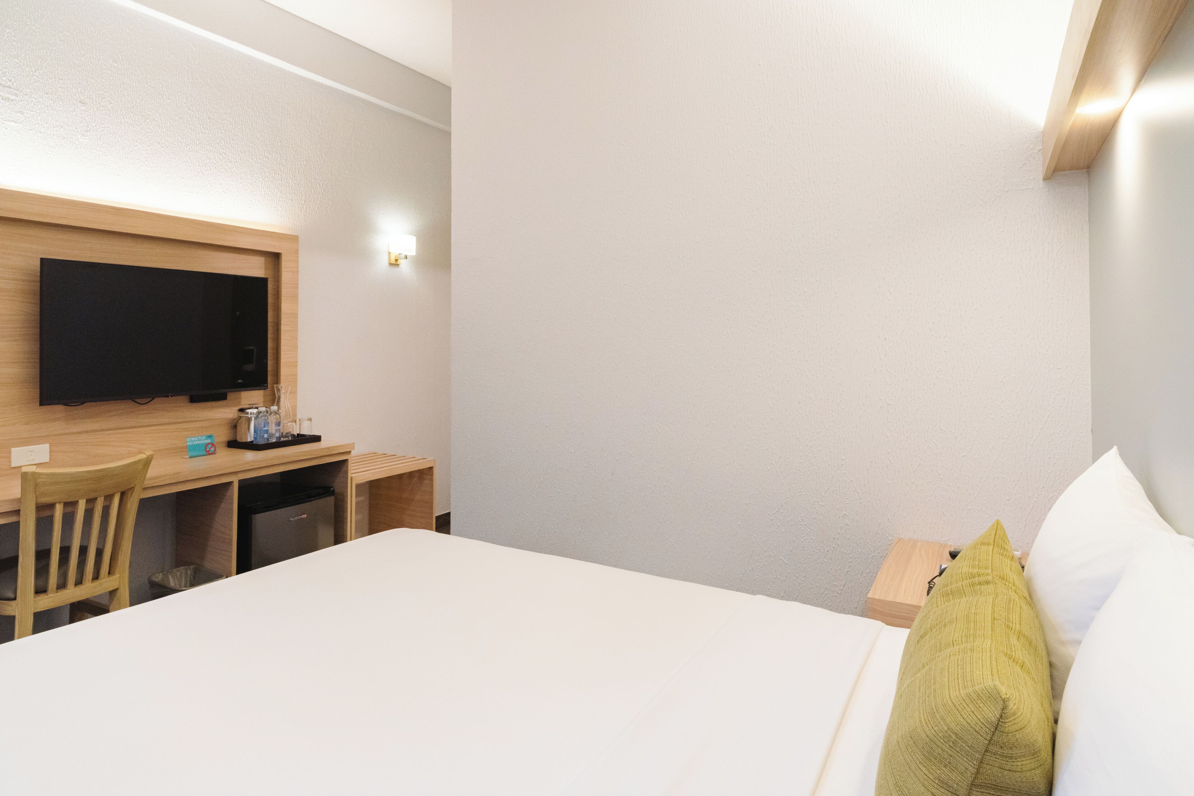 A hotel room with a bed, a desk, a chair, a television, and a small table with a bottle and a box.