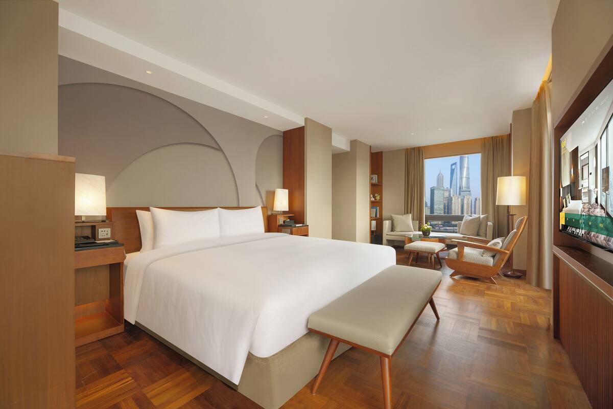 A hotel room with a large bed, a desk, a chair, a television, and a view of a cityscape.
