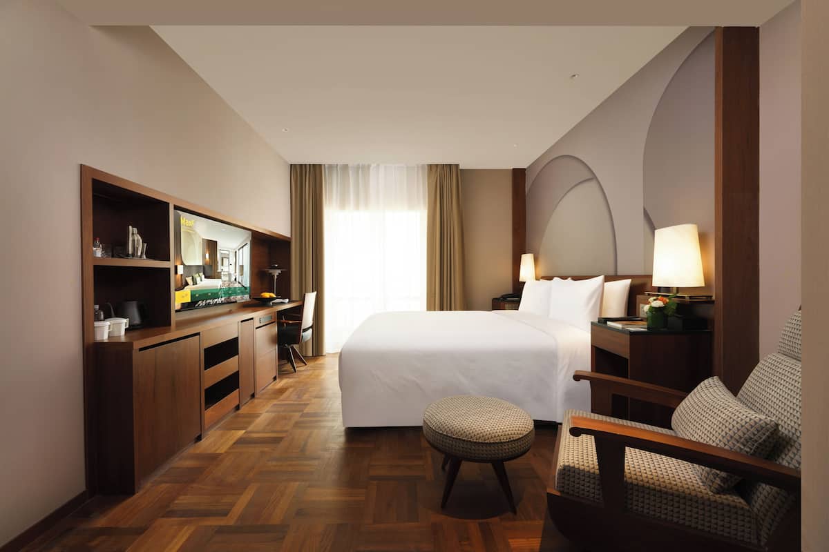 A hotel room with a bed, a desk with a chair, a television, a lamp, and a small table with a flower.