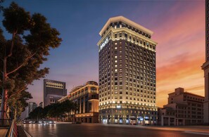 Un hotel de gran altura con un llamativo letrero 'Maxx', rodeado de otros edificios y una calle bordeada de árboles.