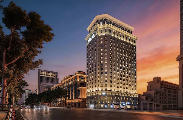 Um hotel moderno e alto com uma placa grande 'Maxx', rodeado por outros edifícios e uma rua com árvores.