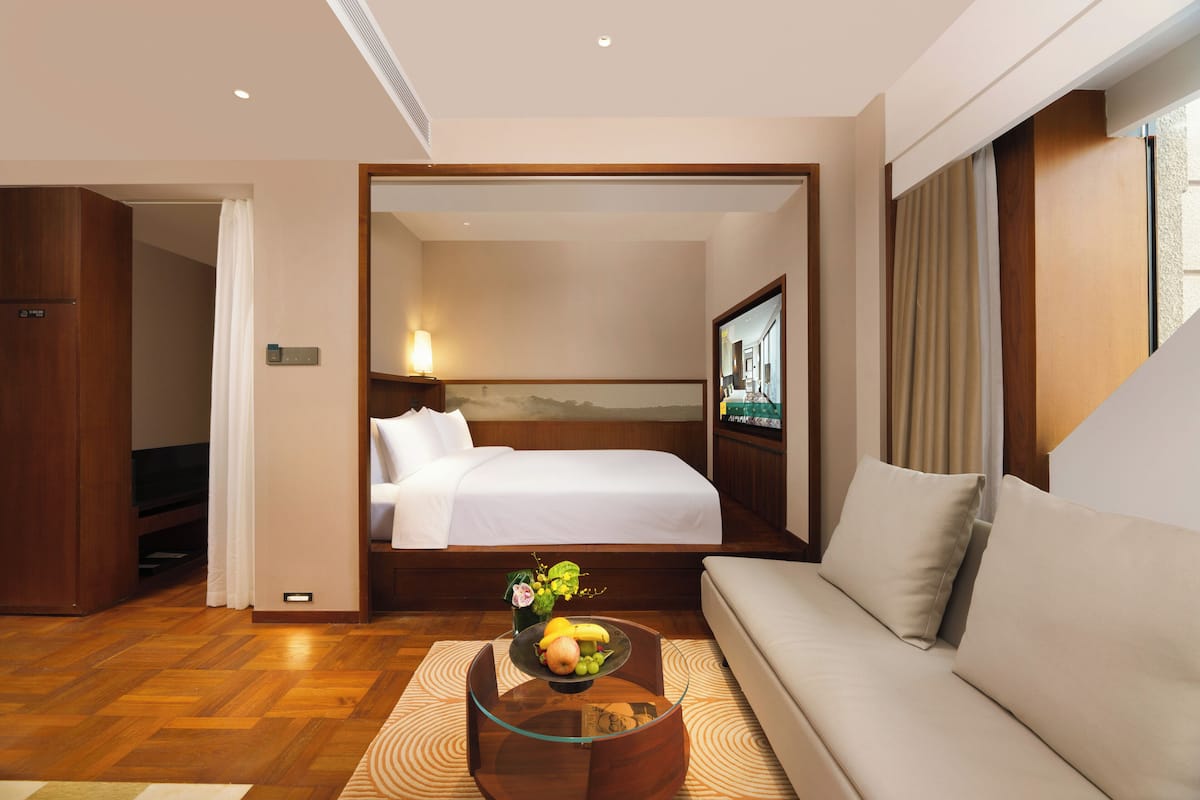 A hotel room with a bed, a sofa, a glass coffee table with fruit, a TV, and a painting on the wall.