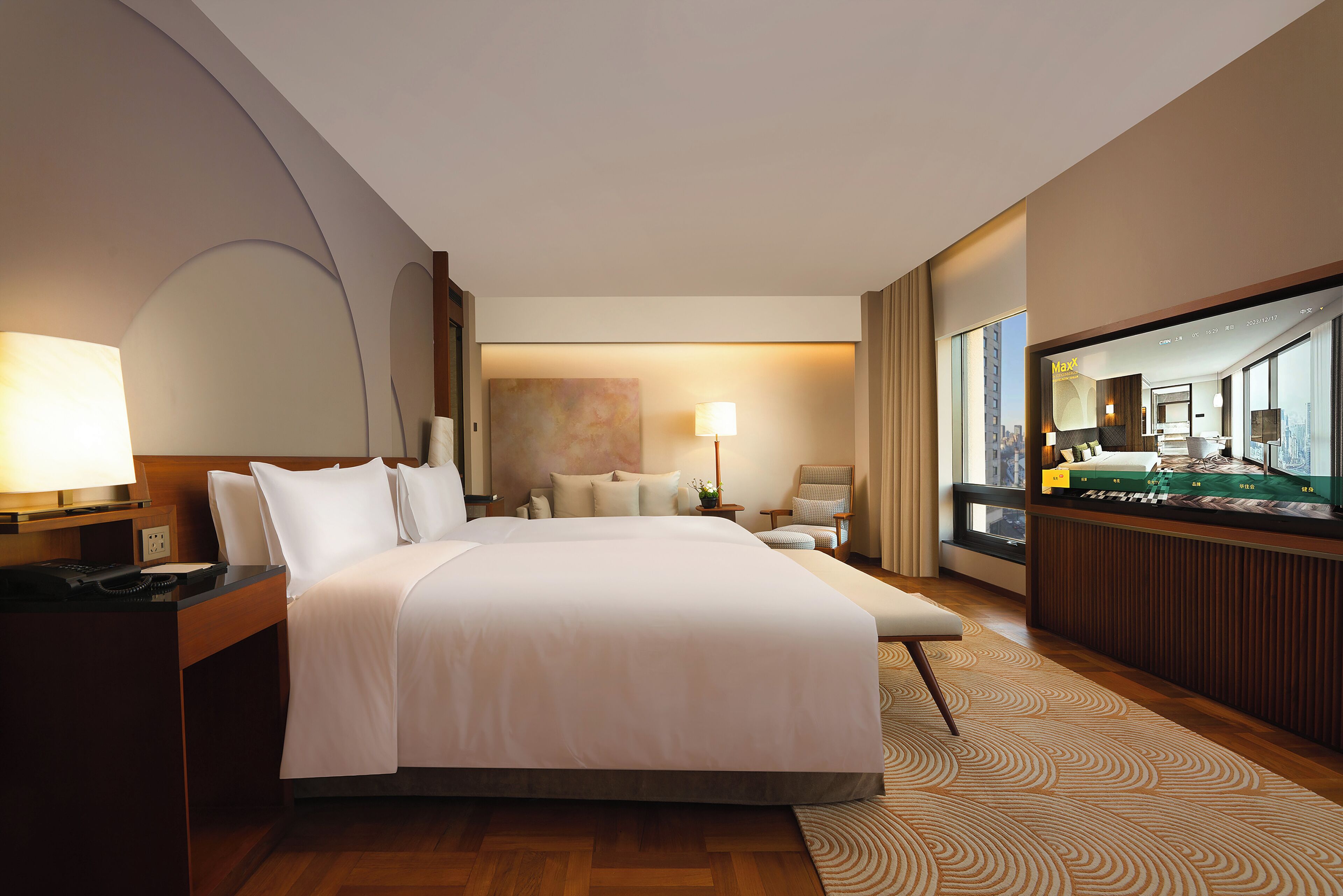 Quarto de hotel com uma cama grande, uma mesa com telefone, uma cadeira, luminária de mesa e um espelho grande, além de vista para a cidade pela janela.