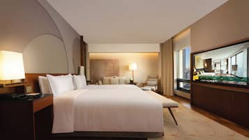 Quarto de hotel com uma cama grande, uma mesa com telefone, uma cadeira, luminária de mesa e um espelho grande, além de vista para a cidade pela janela.