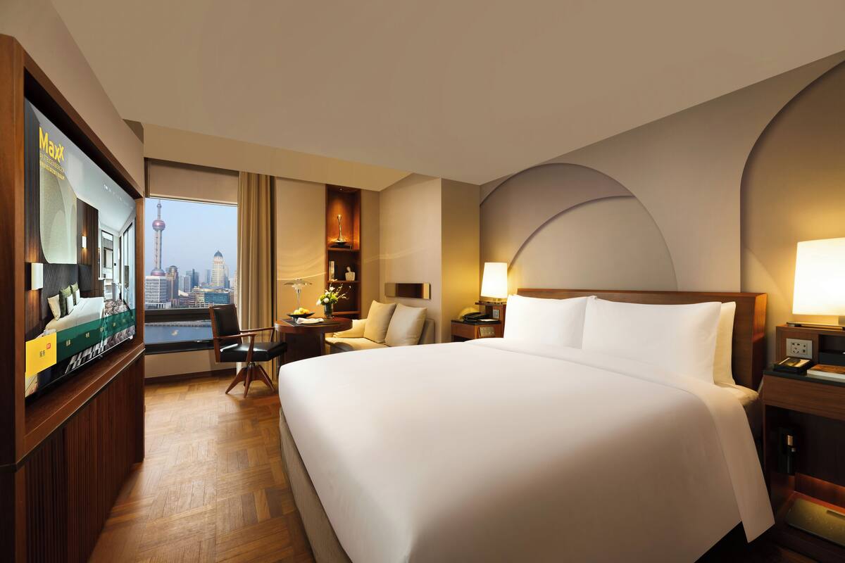 A hotel room with a large bed, a TV, a desk, a chair, a lamp, and a view of a cityscape.