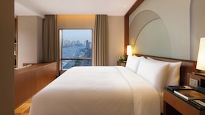 A hotel room with a large bed, a nightstand, a lamp, a view of a cityscape through a window, and a wooden headboard.