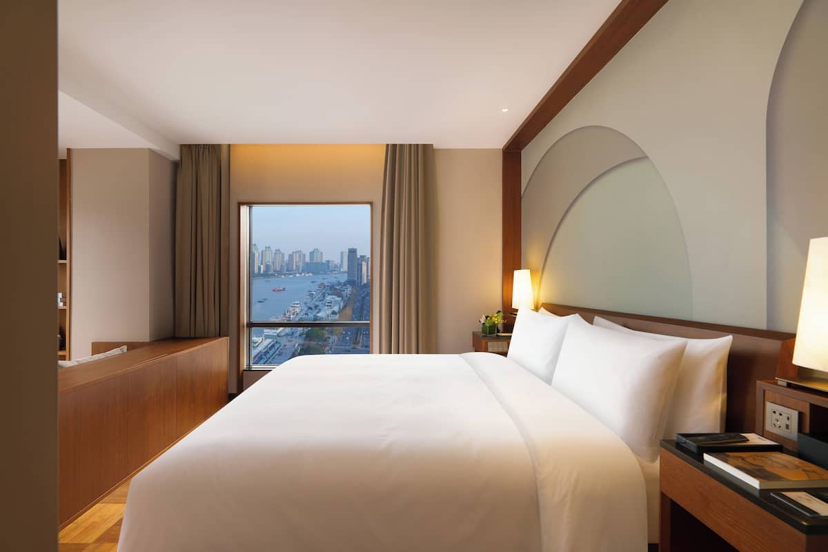 A hotel room with a large bed, a nightstand, a lamp, a view of a cityscape through a window, and a wooden headboard.