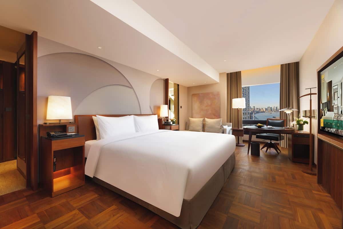 A hotel room with a large bed, a desk, a chair, a small table, a lamp, and a view of a cityscape.