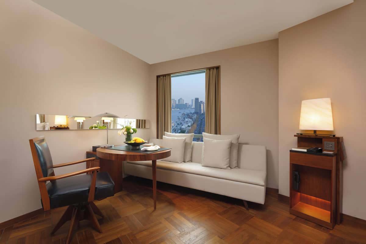 A hotel room with a sofa, a chair, a small table, a lamp, and a view of a cityscape through a window.