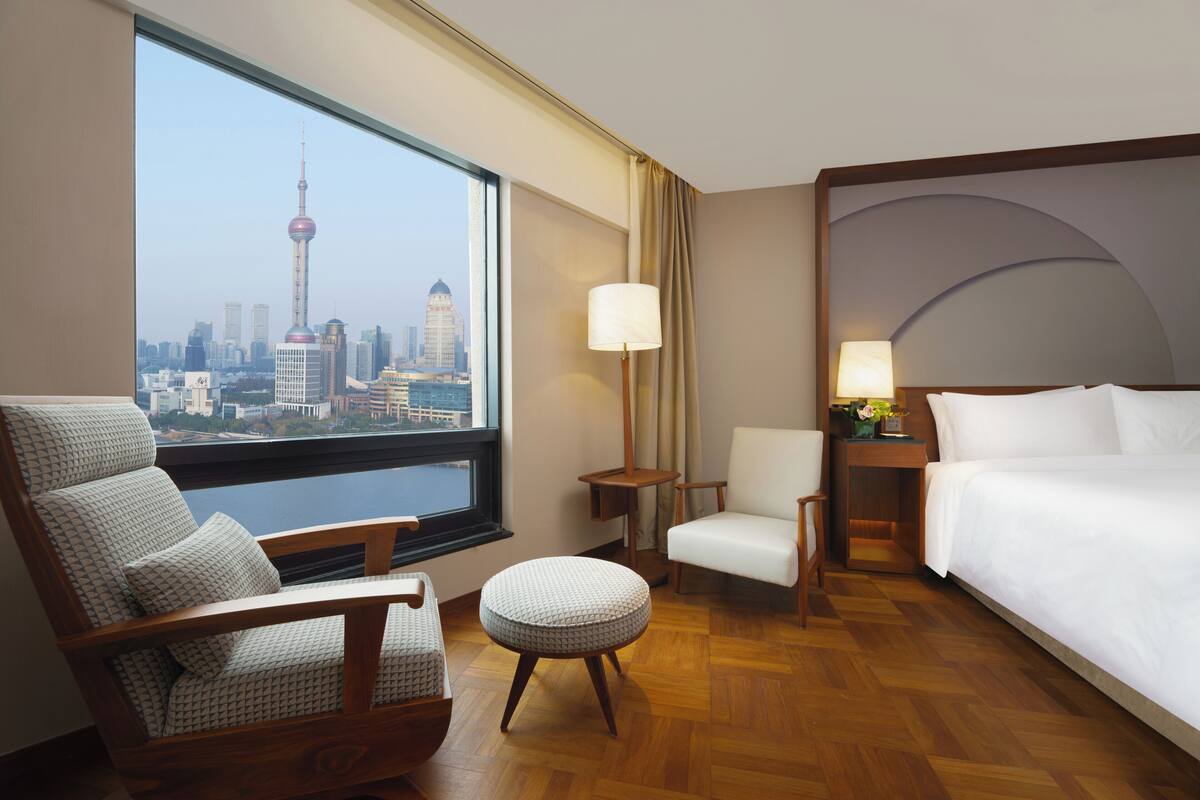 A hotel room with a large window, a bed, a chair, a small table, and a lamp.