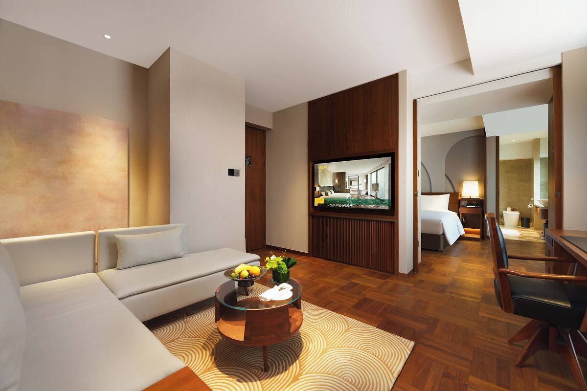 A modern hotel room with a sofa, a coffee table with fruits, a TV, a bed, a desk, and a mirror.