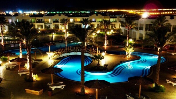 A nighttime view of a resort with a large, illuminated swimming pool, palm trees, and lounge chairs.