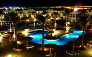 Una vista nocturna de un complejo turístico con una amplia piscina iluminada, palmeras y sillas de descanso.