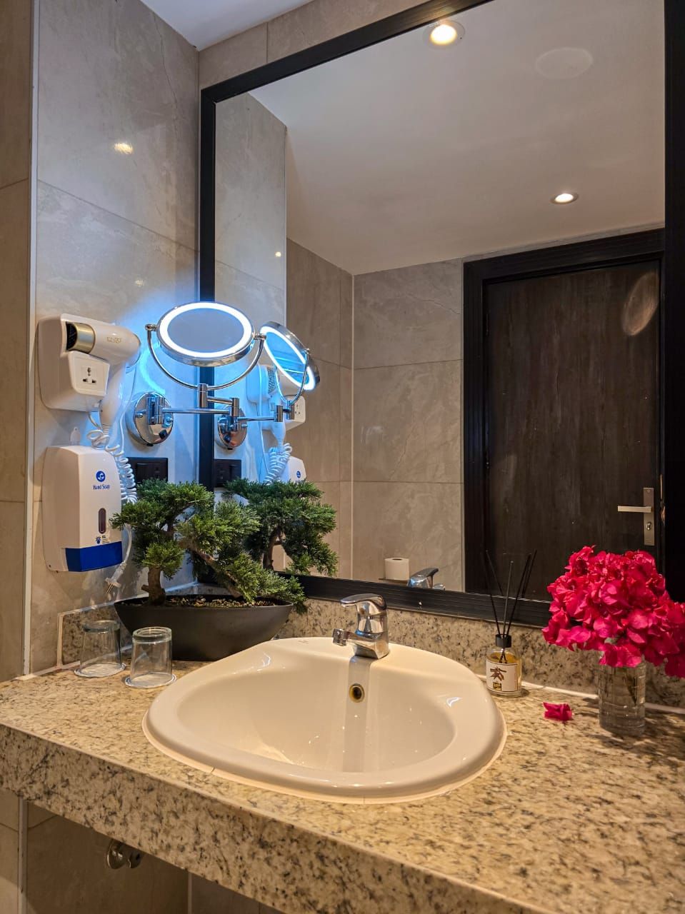 A modern bathroom with a large mirror, a sink, a hairdryer, and a small potted plant.