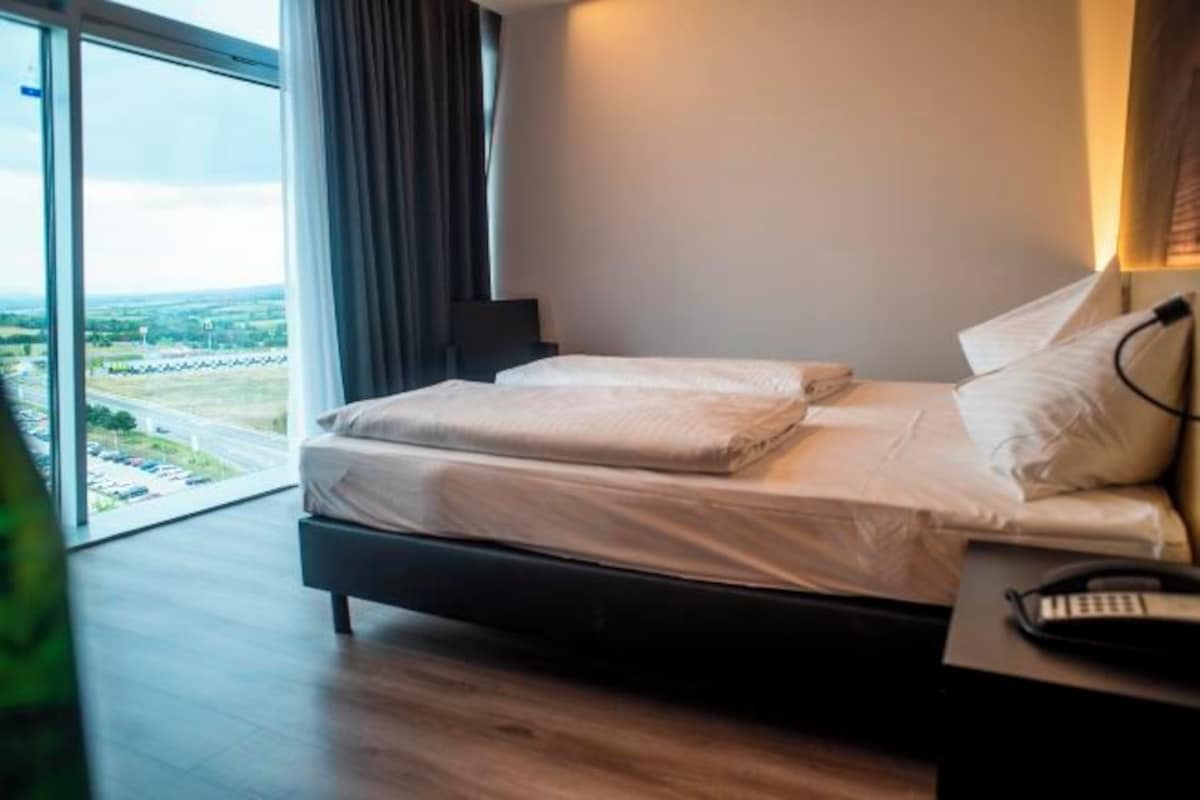 A modern hotel room with a large bed, a bedside table with a phone, and a view of the countryside through large windows.