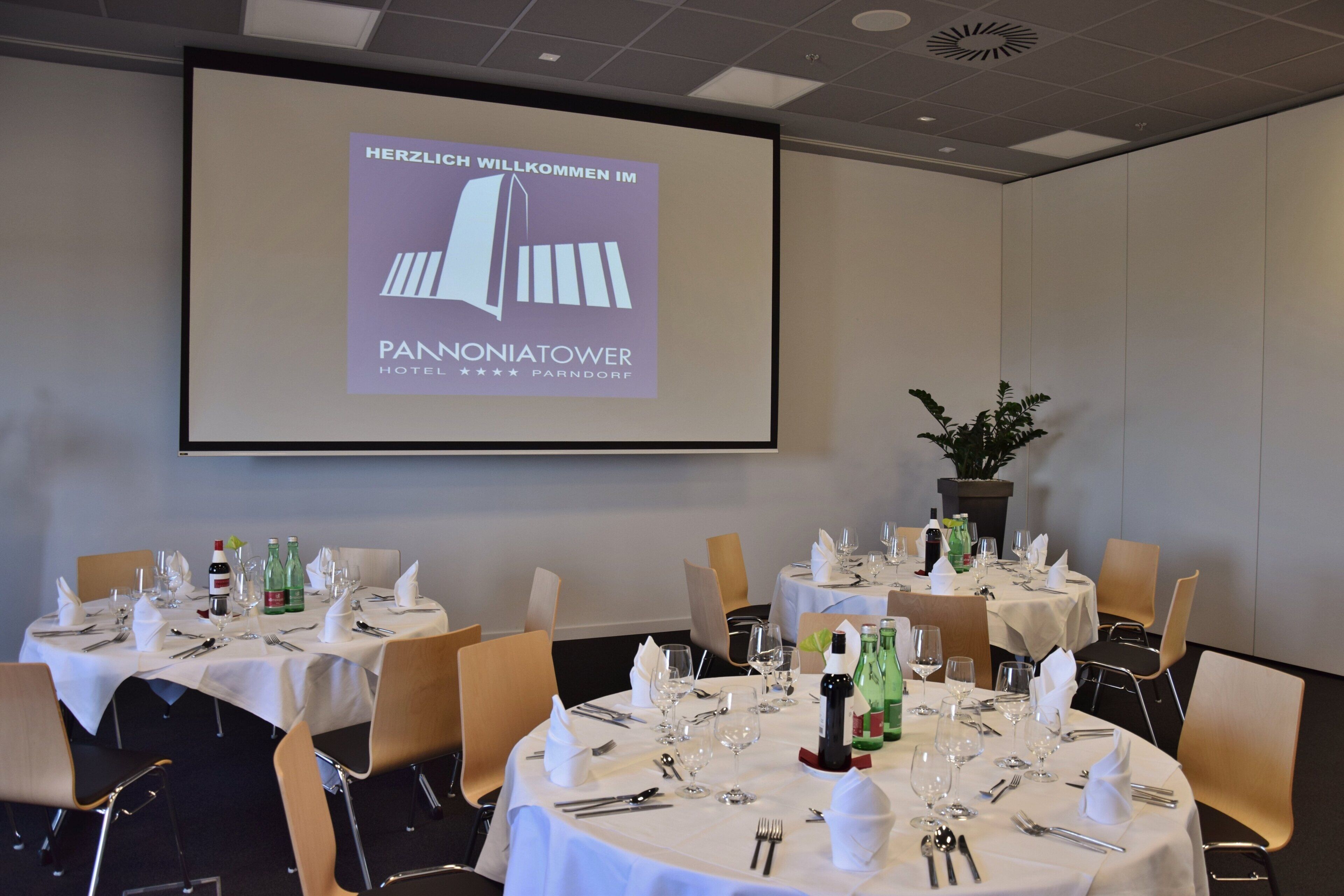 A conference room with tables set for a meeting, a projector screen displaying a welcome message, and a potted plant.