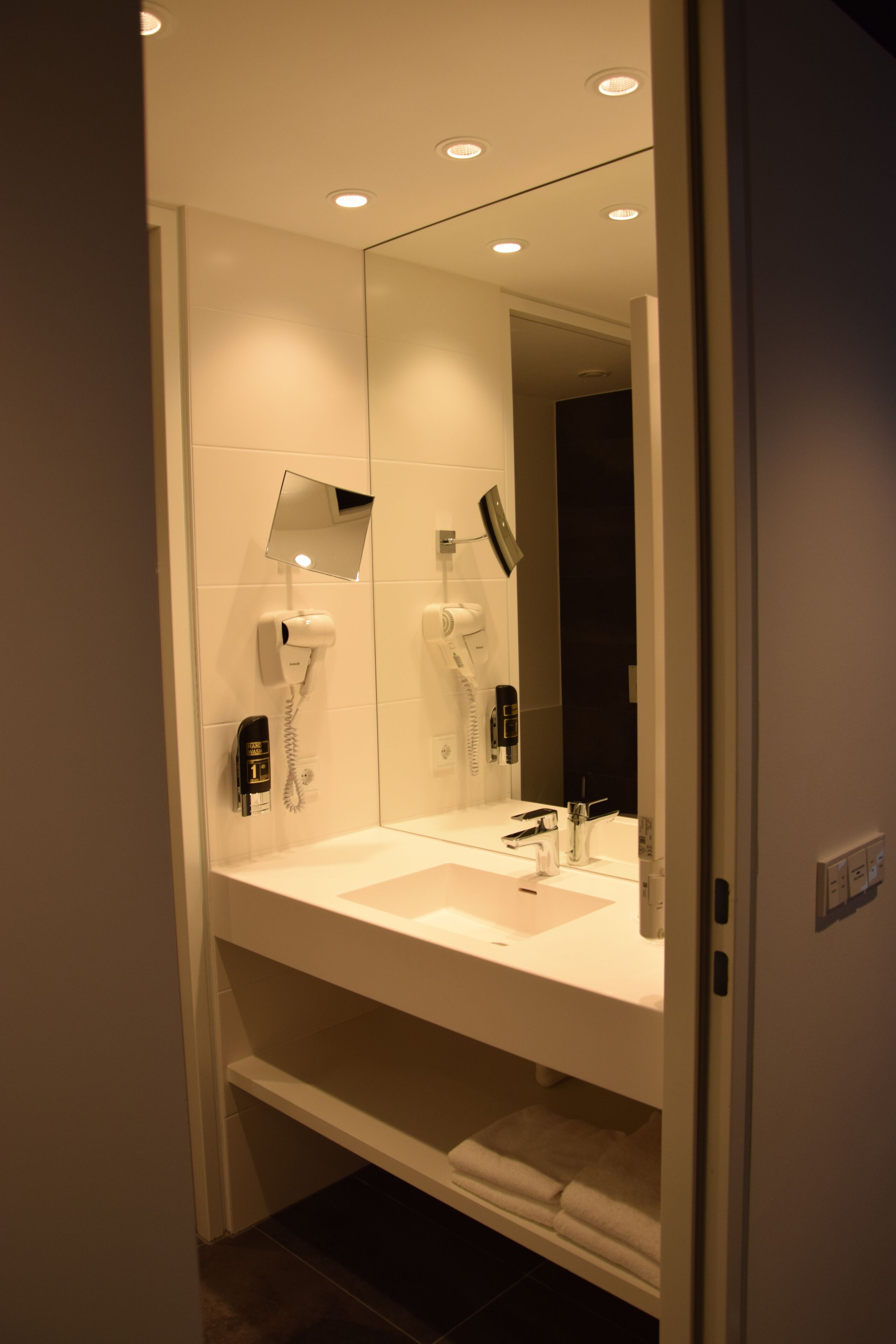A modern bathroom with a white vanity, two hair dryers, a mirror, and recessed ceiling lights.