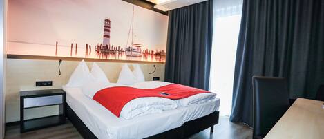 Habitación de hotel moderna con cama, escritorio, mesita de noche y un cuadro grande de una escena costera.
