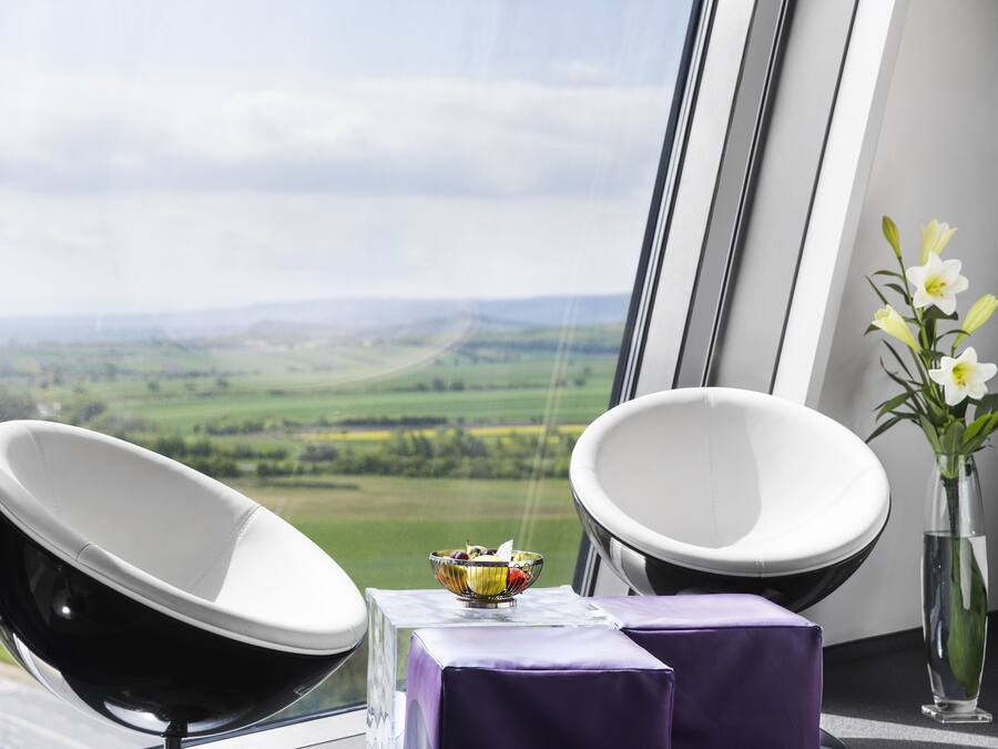 A room with a view of a green landscape, a bowl of fruit on a table, and a vase with flowers.