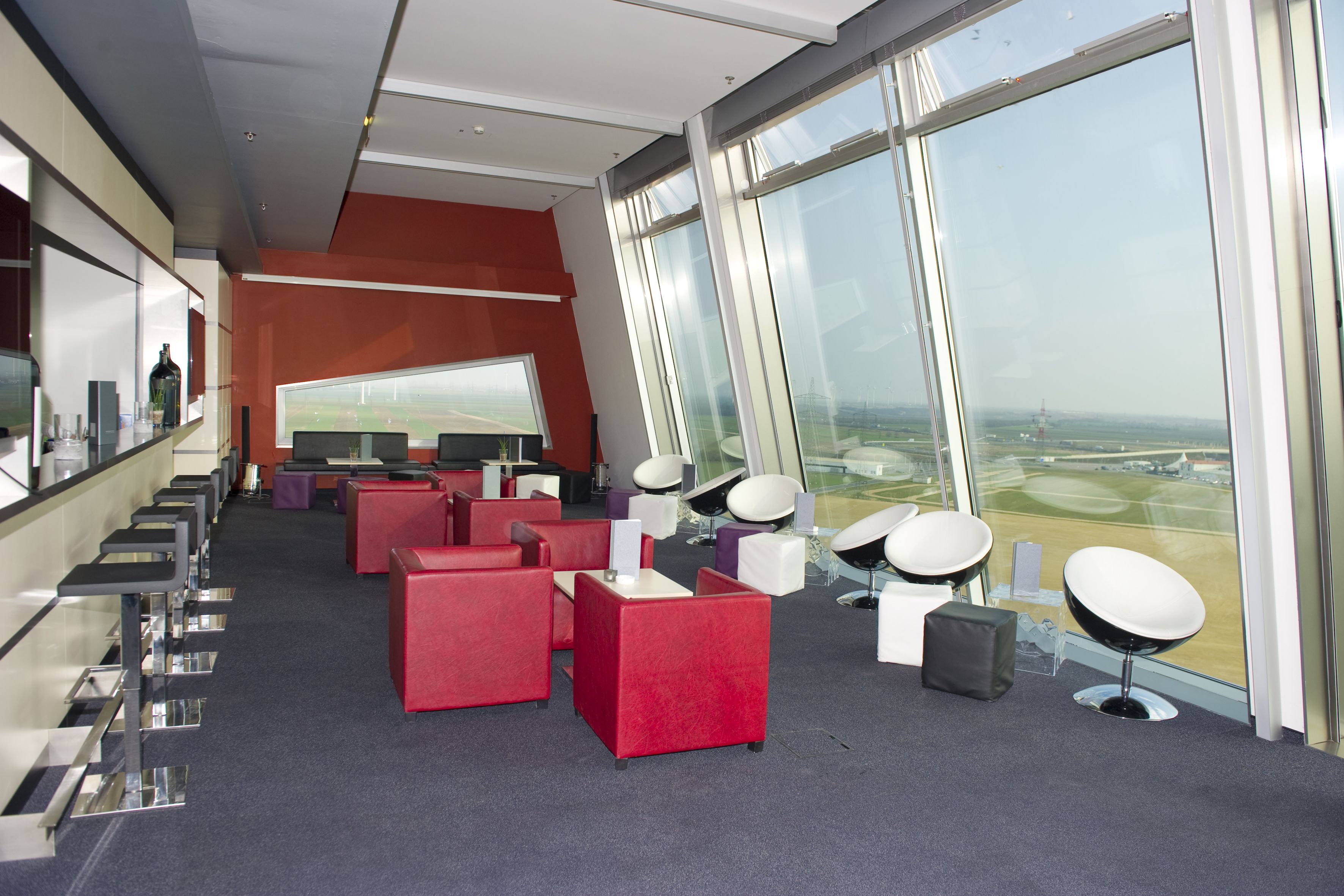 A modern lounge area with red and white seating, a large window offering a view of the airport, and a bar with a glass counter.