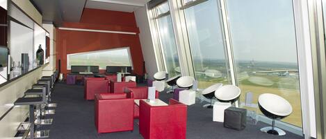 Un área de estar moderna con asientos rojos y blancos, un ventanal con vistas al aeropuerto y una barra con mostrador de vidrio.