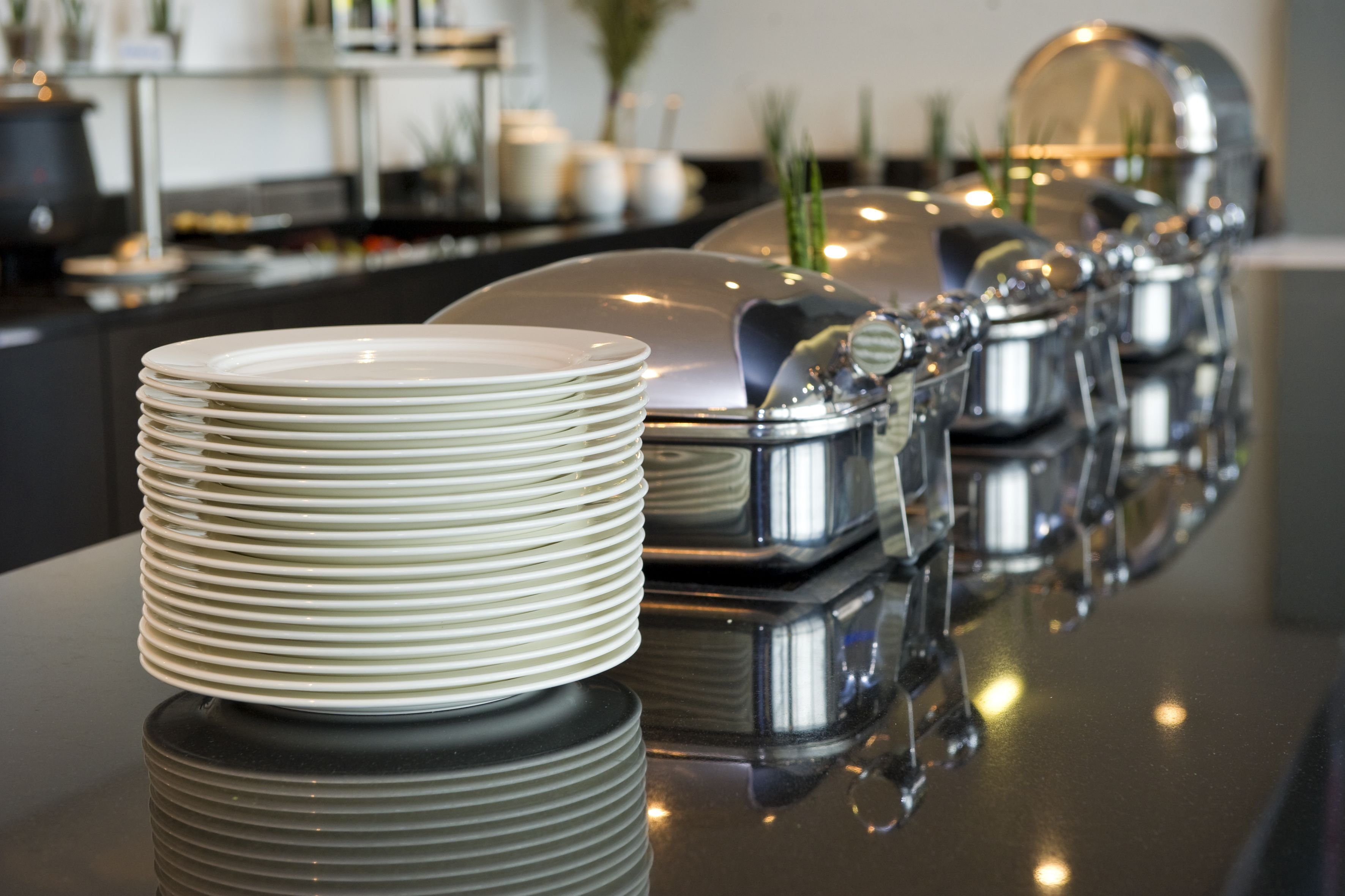 A buffet setup with stacked plates and covered food containers on a reflective surface.