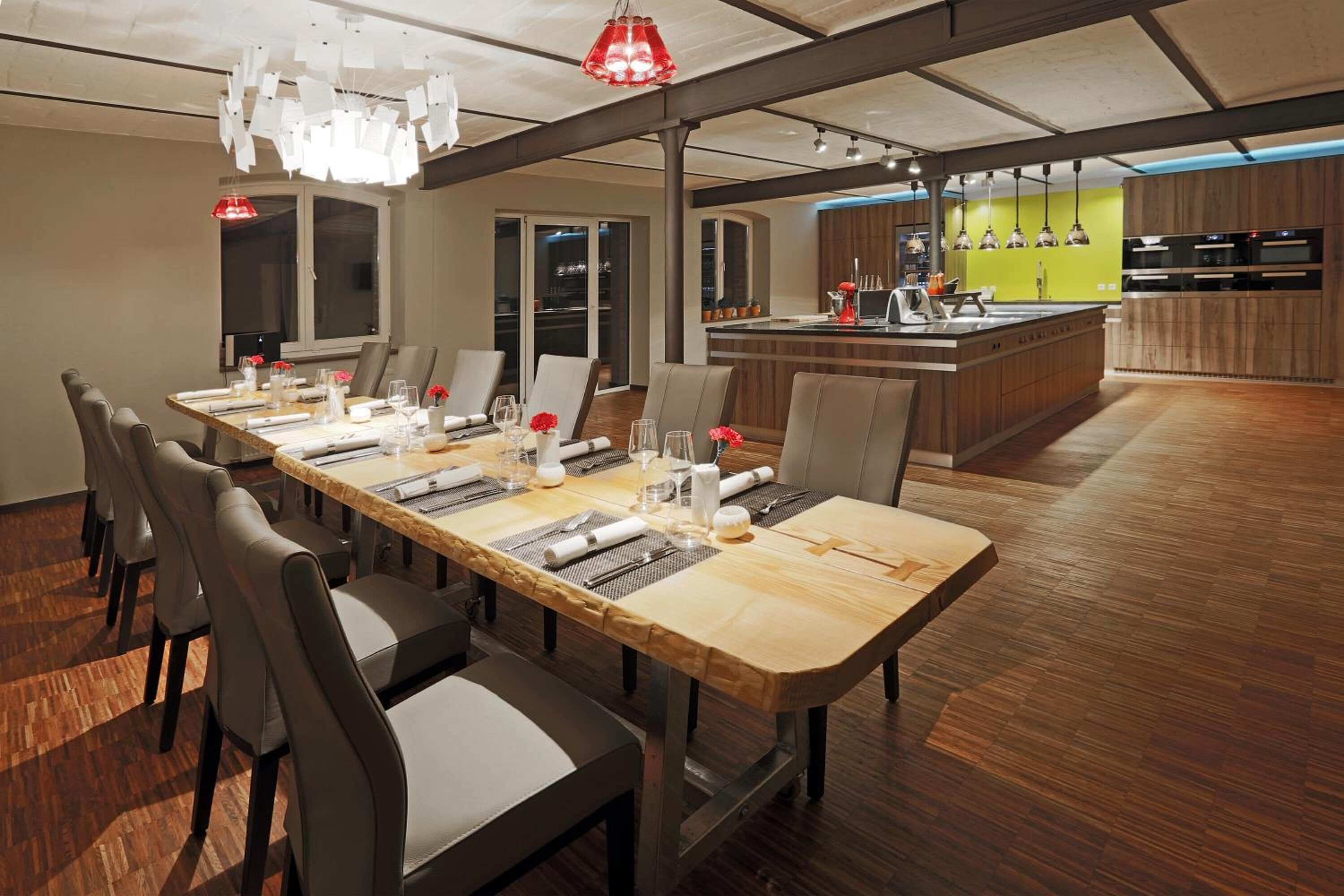 A modern dining area with a long wooden table, multiple chairs, and a kitchen in the background.