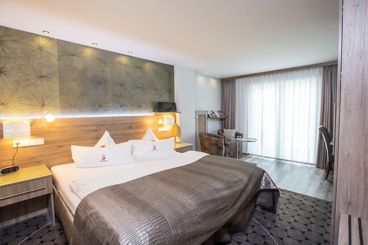 A hotel room with a large bed, a wooden headboard, a bedside table with a lamp, a chair, and a view of the outdoors through a large window.