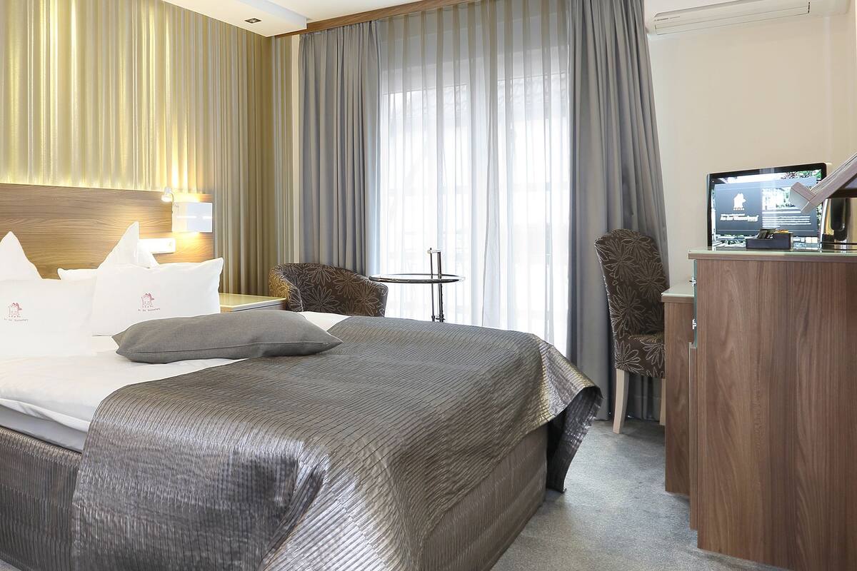 A hotel room with a bed, bedside tables, a desk, a chair, a TV, and curtains.