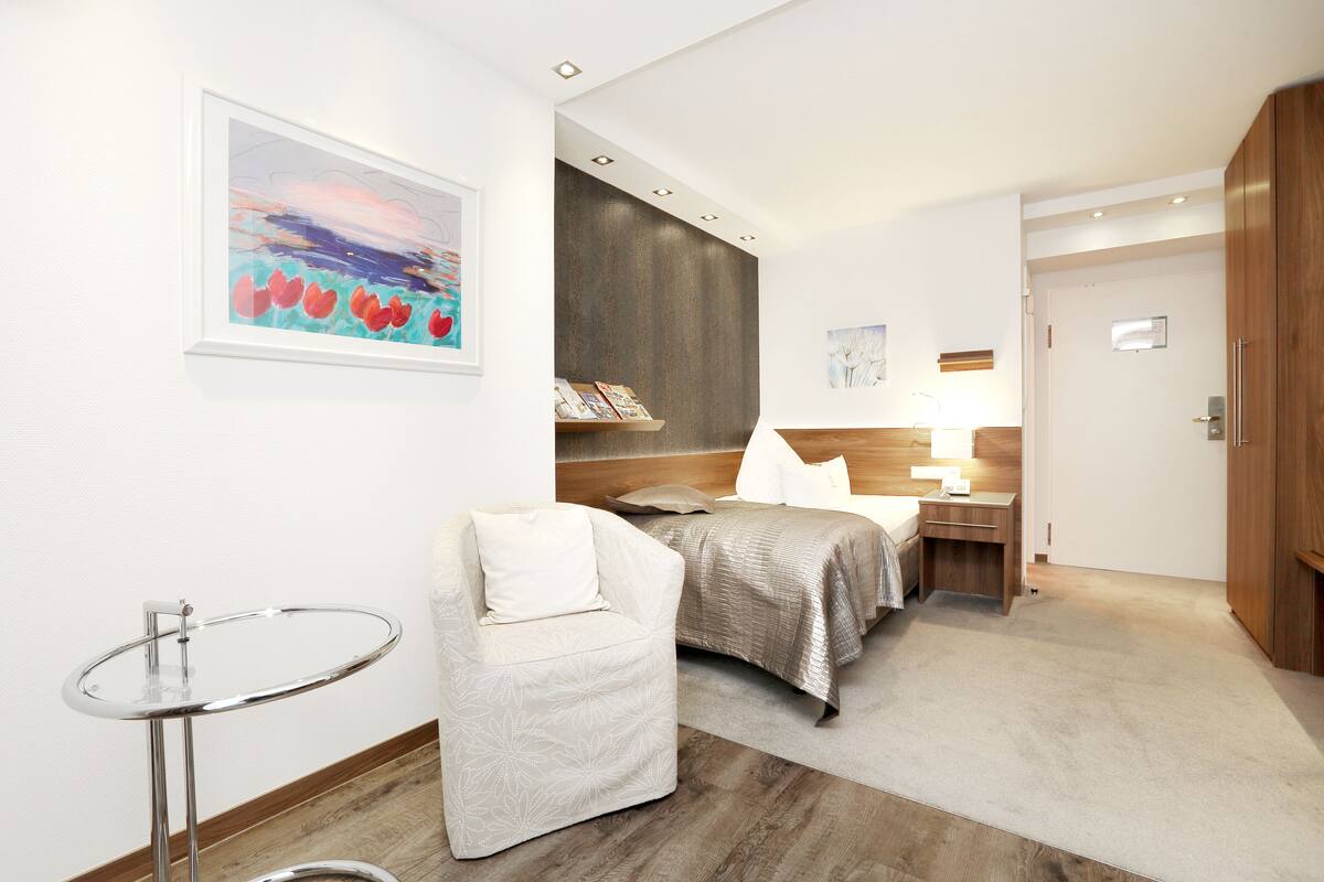 A hotel room with a bed, bedside table, lamp, chair, and artwork on the wall.