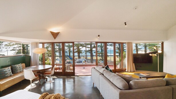 A modern hotel room with a large bed, a grey sofa, a dining area with a table and chairs, and large windows offering a view of the outdoors. - Beach Hotel Resort (Byron Bay)